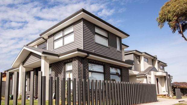 Exterior view of a contemporary multi-storey townhouse with a dark brick ground floor, grey horizontal cladding above, projecting gabled porch roofs, large rectangular windows, and a dark vertical-slat front fence. Similar attached homes continue along the street.