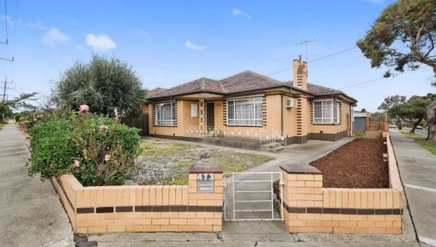 Street-facing view of a single-storey suburban brick bungalow with a low hipped tiled roof, cream brick walls, enclosed front garden, concrete footpaths, mature planting, and a small front entry gate. The home features projecting bay-style window sections, white-framed windows, and a prominent brick chimney.
