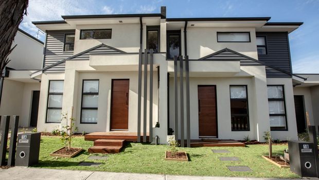 Front exterior view of a contemporary multi-storey attached residential development with two symmetrical entries, white rendered walls, dark horizontal cladding, flat rooflines, timber entry doors, projecting upper-level forms, and narrow landscaped front gardens.