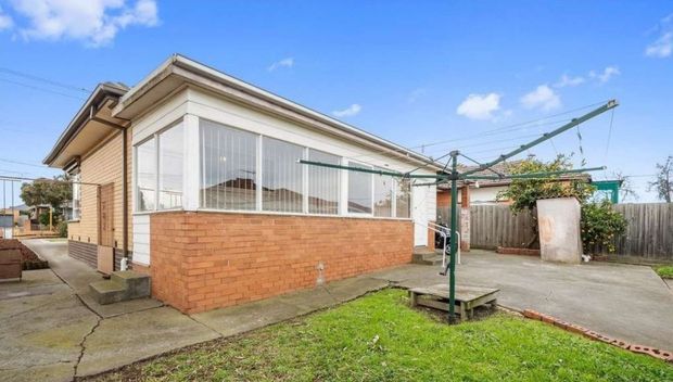 Single-storey suburban brick house viewed from the rear or side, featuring a glazed enclosed sunroom with vertical blinds, a low-pitched hipped roof with broad eaves, concrete paving, a rotary clothesline, and a small lawn enclosed by timber fencing.
