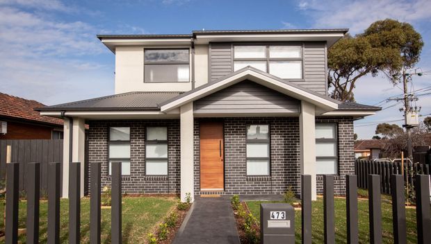 Front exterior view of a contemporary two-storey suburban residence with a dark brick lower facade, light grey horizontal cladding and rendered upper walls. The home has a prominent front-facing gable, low-pitched metal roof, timber entry door, vertical dark fence and a paved path leading to the front door.