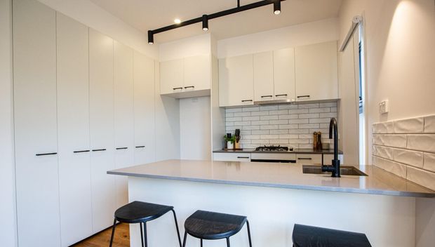 Bright, compact contemporary kitchen with extensive white flat-panel cabinetry, a pale stone island bench with integrated sink and black tapware, subway-tile splashback, timber flooring, and black track lighting. Three black stools sit at the island, and a textured tiled wall is visible at right.