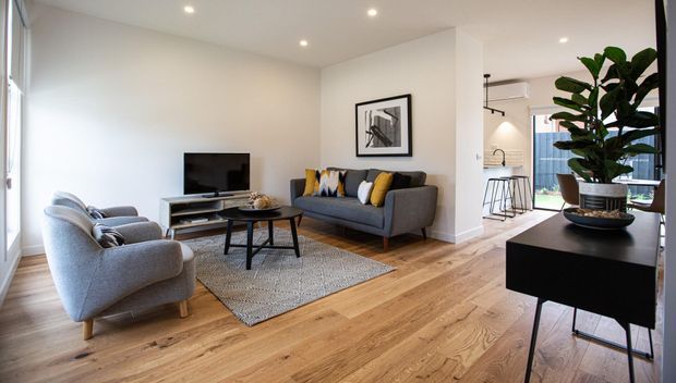 A bright, contemporary open-plan living room with a grey sofa and matching armchairs arranged around a black coffee table and rug. Light timber flooring, white walls, recessed ceiling lighting, framed artwork, and a black console with a large indoor plant create a clean, warm interior. A kitchen/dining area is visible beyond the living space.