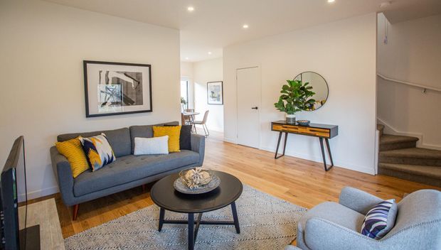 A bright, contemporary living room with a charcoal-grey sofa, light blue armchair, black round coffee table, patterned rug, timber flooring, and mustard and blue accent cushions. A slim console with a round wall mirror sits against the white wall, while a carpeted staircase and hallway are visible to the right and rear.