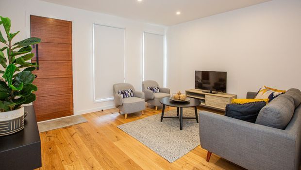 Bright, compact contemporary living room with warm timber flooring, a large timber entry door, neutral walls, two armchairs, a grey sectional sofa, a round black coffee table, a low media console and television. White roller blinds, recessed ceiling lights and a large indoor plant complete the clean, inviting space.