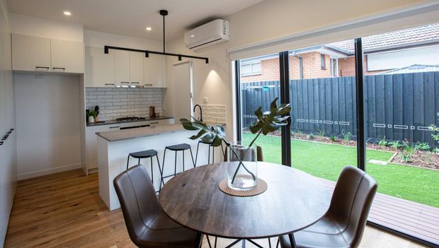 Bright open-plan kitchen and dining area with light cabinetry, white subway-tile splashback, timber flooring, a peninsula with bar stools, and a round timber dining table beside large sliding glass doors. The glazing opens to a small landscaped backyard with decking and lawn. Contemporary black-framed lighting and window detailing provide contrast against the neutral interior.