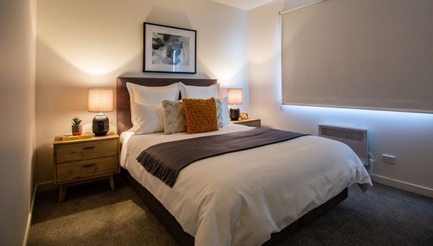 A compact, warmly lit bedroom with a neatly made bed, upholstered dark headboard, white bedding, textured mustard and neutral accent cushions, and a dark throw. Matching bedside lamps sit on timber bedside tables, with framed artwork above the bed. The room has neutral painted walls, carpet flooring, and a large window fitted with a grey roller blind.