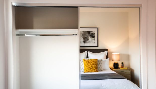 A built-in sliding-door wardrobe is shown open, with an empty hanging rail and upper storage shelf on the left. Through the opening, a neatly styled bedroom is visible with a bed, bedside table, lamp, framed artwork, and warm neutral finishes.