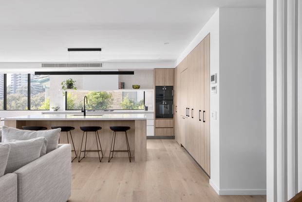 Bright, open-plan contemporary kitchen viewed from an adjacent living area. The kitchen features a long light-wood island with a dark stone-look benchtop, three bar stools, integrated appliances, extensive light timber cabinetry, black-framed glazing, and pale timber flooring. A sofa is visible in the foreground, with urban buildings and greenery seen outside.