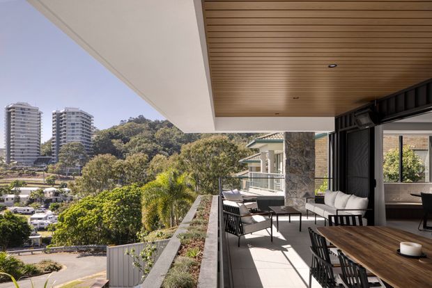 A spacious covered outdoor terrace or balcony adjoining a contemporary residence, furnished with an outdoor dining table and chairs. The terrace has a warm timber slatted ceiling, pale tiled flooring, dark-framed full-height glazing, stone-clad columns, built-in planter beds, and views toward a leafy urban hillside with high-rise buildings.