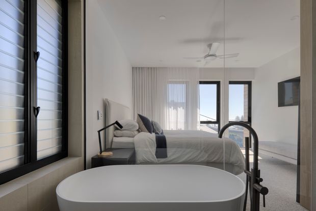 A bright, minimalist bedroom with an integrated freestanding bathtub in the foreground. The room features a low upholstered bed, neutral bedding, a dark bedside table and angled task lamp, full-height curtains, tall black-framed glazing, a ceiling fan, and a wall-mounted television. A compact louvered window is visible on the left, with an open-plan bathroom zone suggested by the bathtub and shower fitting.
