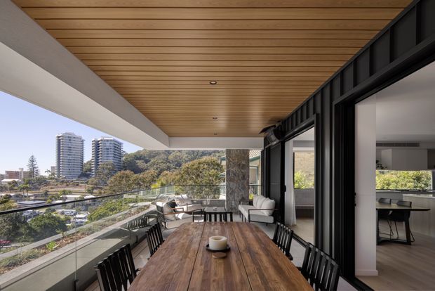 A spacious covered balcony terrace with a long solid timber dining table and black dining chairs, opening directly into a contemporary interior through large black-framed sliding glass doors. The terrace has a timber-lined ceiling, glass balustrade, exposed stone-clad column, outdoor lounge seating, and elevated urban views across landscaped surroundings and apartment buildings.