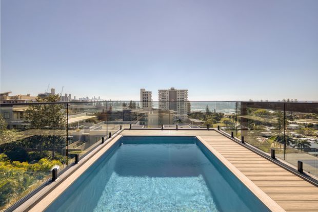 Rooftop or elevated terrace swimming pool with a timber-look deck and clear glass balustrades, overlooking a coastal urban landscape and the sea. The image is taken outdoors in bright daylight, with modern apartment buildings and landscaped surroundings visible beyond the pool.