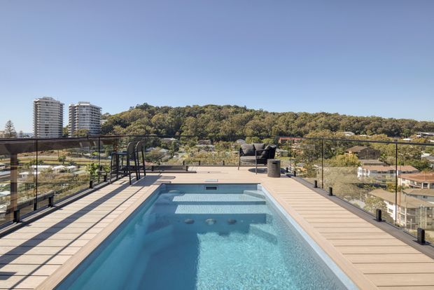 Rooftop or elevated outdoor terrace with a rectangular swimming pool, timber-look decking, glass balustrades, outdoor seating, and a compact dining setting. The terrace overlooks a leafy suburban or urban hillside with apartment buildings in the distance under a clear blue sky.