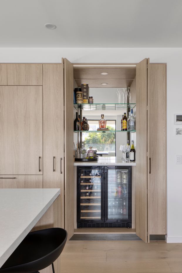 A built-in drinks station within a contemporary kitchen, featuring full-height light timber cabinetry, open glass shelving displaying liquor and glassware, and an under-counter wine and beverage refrigerator. Part of a pale stone kitchen island and a black bar stool are visible in the foreground. The space is bright and clean with warm wood tones and a view through the shelving to greenery outside.