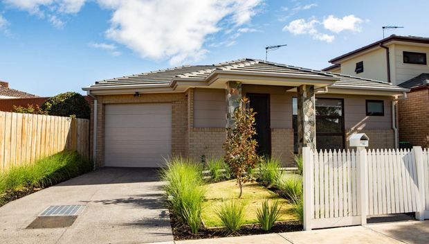 Street-facing view of a single-storey suburban brick house with a low-pitched tiled hip roof, enclosed single garage, covered front porch supported by stone-clad columns, and a small landscaped front garden behind a white picket fence and gate.
