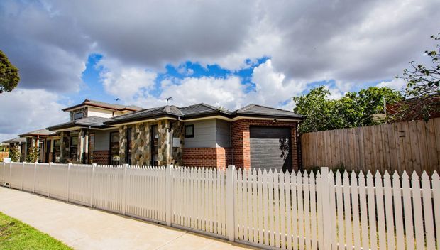 Single-storey suburban residential home viewed from the street, with a low-pitched tiled roof, brick and stone-clad facade, covered front porch, garage, timber boundary fencing, and a white picket fence along the sidewalk. The house has a varied roofline and a warm, established streetscape setting.