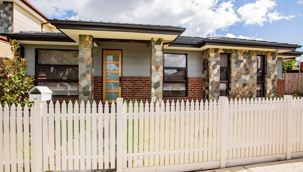 Single-storey residential house facade viewed from the street, featuring a low-pitched dark roof, pale horizontal cladding, varied natural stone wall sections and columns, red brick accents, timber-framed glazed entry door, and a white picket fence in the foreground.