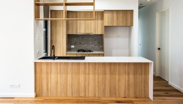 Compact contemporary kitchen with a timber-grain island and integrated cabinetry, a white stone-look benchtop, dark tiled splashback, black tapware and cooktop, open shelving, and timber flooring. A hallway and interior doorway are visible to the right.