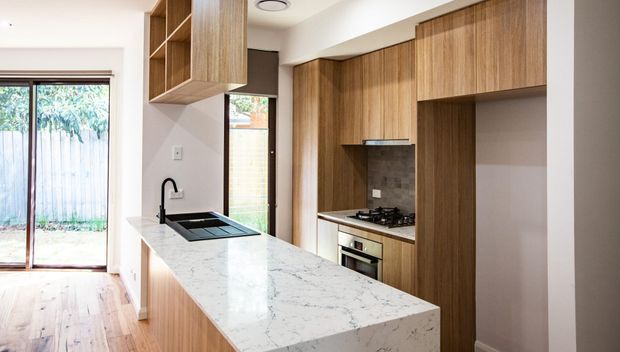 A compact contemporary kitchen with warm timber cabinetry, a white veined stone island bench, integrated cooktop and oven, and a dark tiled splashback. A large glazed door brings in natural light and provides a view to an outdoor area. Timber flooring and minimal white walls create a warm, clean interior.