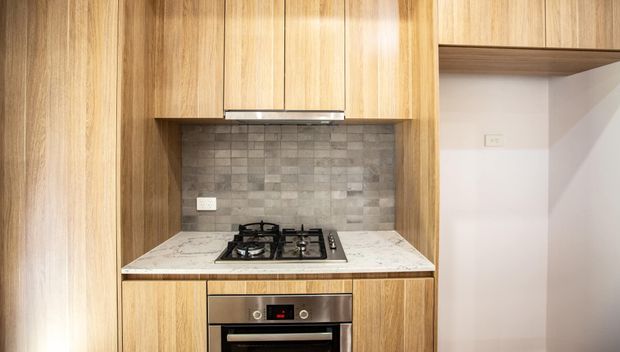 Compact contemporary kitchen with warm timber cabinetry, a built-in gas cooktop and oven, a light stone-look benchtop, and a grey textured tiled splashback. The cabinetry extends to the ceiling, with an open shelving recess visible on the right.