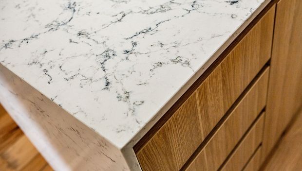Close-up detail of a white veined stone benchtop with a thick waterfall edge meeting warm timber cabinetry with recessed drawer fronts. The image focuses on the joinery and countertop materials rather than a complete room.