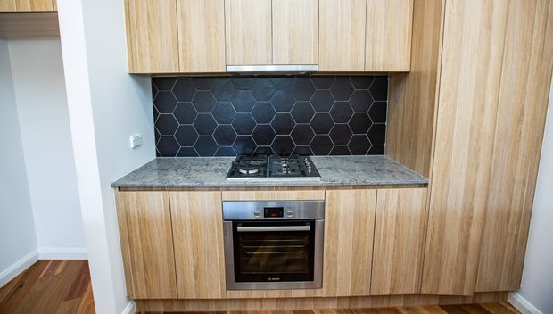Compact modern kitchen with full-height light timber cabinetry, a dark hexagonal tile splashback, grey stone benchtop, integrated gas cooktop and built-in oven. Dark timber flooring and white walls complete the clean, contemporary fit-out.