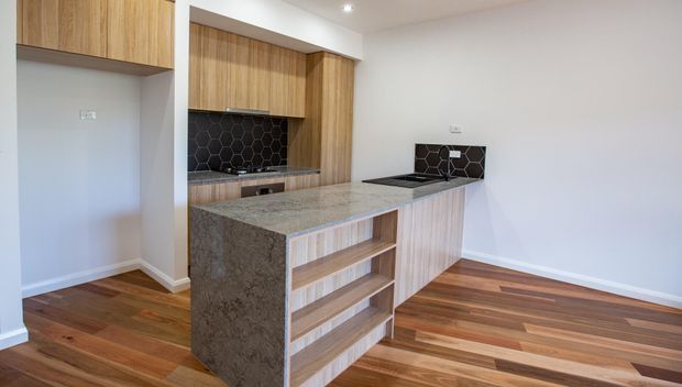 Contemporary kitchen with a large central island, timber cabinetry, open shelving, dark hexagonal tile splashbacks, a stone benchtop, and warm timber flooring. The bright space has white walls and recessed ceiling lighting.