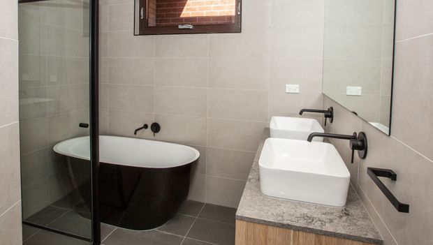 Modern bathroom featuring a freestanding oval bathtub beside a frameless glass shower enclosure, double vessel basins on a timber vanity with a grey stone benchtop, dark wall-mounted tapware, and large-format grey tiled walls and floor. A small high window provides natural light.