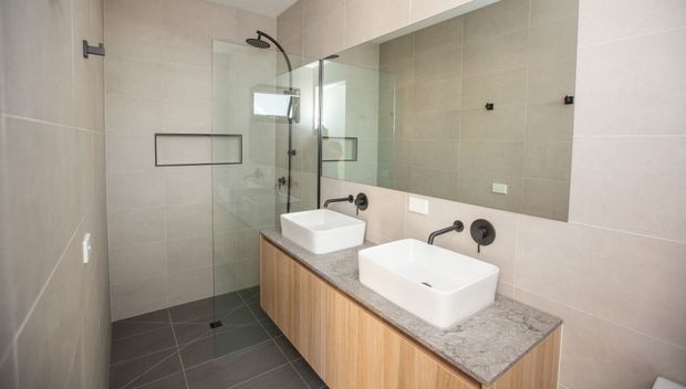 Contemporary double-vanity bathroom with a walk-in glass shower. Large-format pale beige wall tiles and dark grey floor tiles create a neutral contrast, while a timber vanity with a speckled stone benchtop supports two white countertop basins. Matte black wall-mounted tapware and shower fittings provide a restrained modern accent.