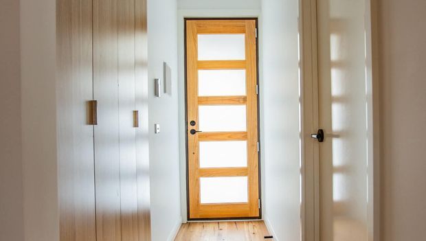 Bright, narrow entry hall featuring a contemporary timber front door with multiple horizontal glazed panels, pale walls, light timber flooring, and full-height built-in cabinetry with simple dark pulls along one side. A white interior door opens on the opposite side.