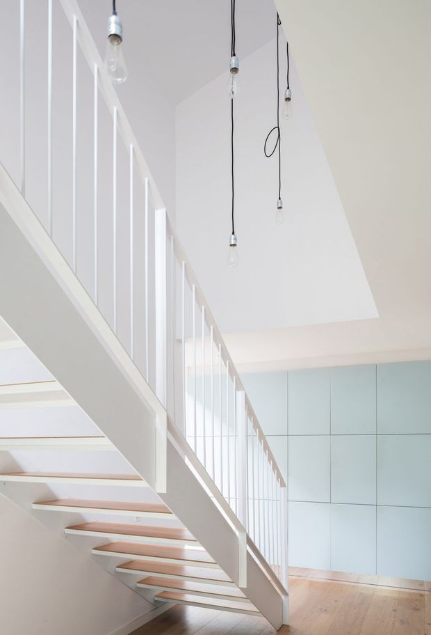 Bright minimalist interior stairway with an open-riser staircase, white structural stringers and slender vertical balustrade, pale timber stair treads, light timber flooring, and a large white geometric ceiling plane. Several exposed pendant bulbs hang from black cords, while pale blue-grey full-height wall panelling forms the background.