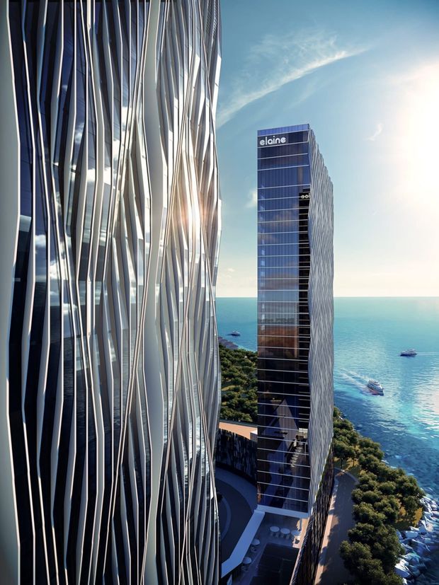 A high-angle architectural exterior view of two tall contemporary glass towers beside a bright blue coastline. The foreground tower has a highly distinctive facade of irregular, vertically undulating metallic fins, while the slimmer background tower has dark blue glazed curtain walls with similar vertical side articulation and visible signage. Landscaped grounds, a rooftop or podium area, and boats on the water are visible below and beyond the buildings.