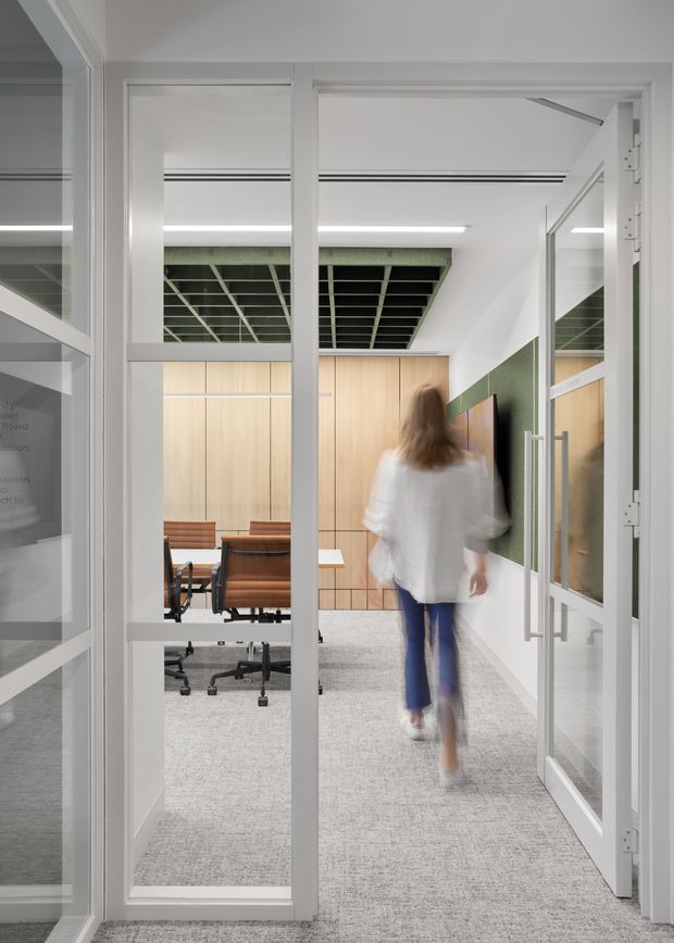 A bright contemporary office interior viewed through glazed framed doors. The corridor has light grey carpet, white walls and doors, pale timber wall panelling, green acoustic ceiling panels and a matching green wall feature. A meeting room with a timber table and office chairs is visible to the left, while a blurred person walks through the corridor.