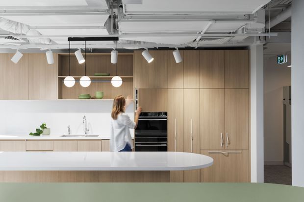 Contemporary open-plan kitchen with extensive light timber cabinetry, integrated black appliances, a white island bench with a rounded end, and a white splashback. Open shelving with restrained decor sits beneath a linear track of pendant and spot lighting. The exposed white ceiling services and ductwork give the space a refined commercial or office fit-out character.