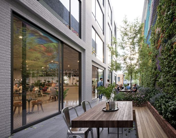 A contemporary urban outdoor dining terrace beside a modern multi-storey building. A long timber dining table with metal chairs sits on grey paving, bordered by built-in timber seating and dense vertical greenery. Full-height glazed doors reveal a casual café or dining interior, while pedestrians and planting occupy the narrow landscaped courtyard-like space.