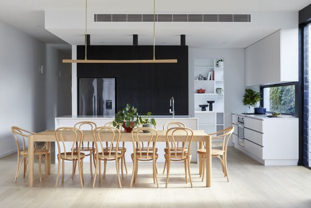 Bright open-plan contemporary kitchen and dining area with a large light timber dining table and matching bentwood chairs in the foreground. The kitchen features white cabinetry, a dark feature wall with integrated stainless-steel refrigerator, white open shelving, a pale benchtop, pendant lighting, and large windows overlooking greenery. Light timber flooring and a restrained neutral palette create an airy, refined interior.
