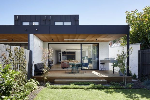 Modern single-storey rear extension opening onto a landscaped garden, with a broad flat roof, dark vertical cladding, white brick walls, warm timber soffit and decking, and large sliding glass doors. The covered outdoor area includes lounge and dining furniture plus an outdoor barbecue or kitchen zone.