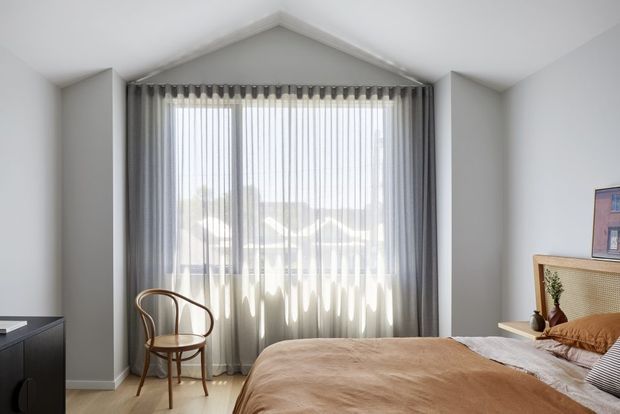 Bright contemporary bedroom with a peaked ceiling detail, large window dressed in sheer and dark grey curtains, a warm upholstered bed, cane bedside furniture, and a bentwood chair. The room has a restrained neutral palette with natural timber accents and abundant daylight.