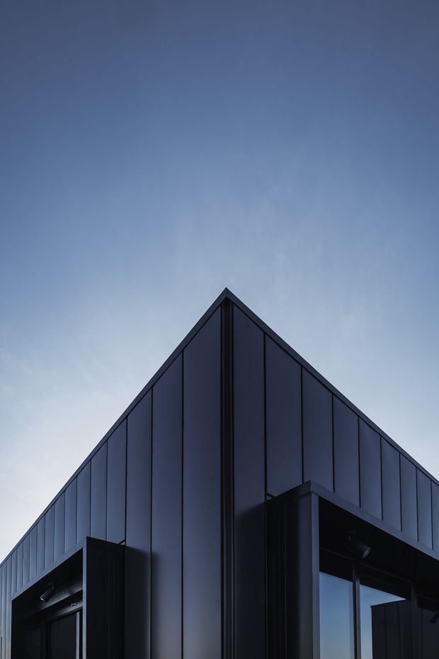 Close-up upward view of a contemporary building corner with a sharply peaked gable roofline, dark vertical metal cladding, black-framed glazing, and strong geometric facade detailing against a blue sky.