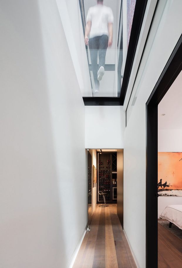 A narrow, minimalist interior hallway with white walls and warm timber flooring. A large overhead glazed skylight reveals a person walking above, creating a dramatic vertical connection between levels. At the end of the corridor is a compact dark built-in wine storage and bar area, while an adjacent bedroom is partially visible through a black-framed opening on the right.