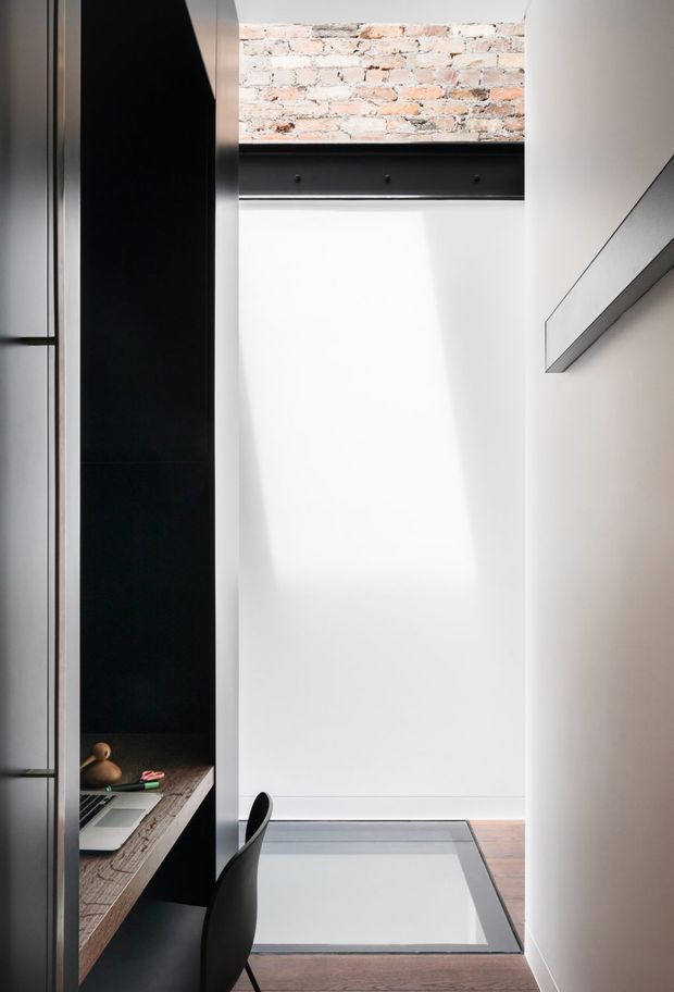 A narrow, vertically oriented home office or study nook with a built-in dark storage and desk unit along the left wall. The space has a minimalist white wall, exposed brickwork and a black steel beam near the ceiling, a slim projecting shelf or cabinet on the right, a black desk chair, and a glazed floor inset or skylight-like feature set into the timber floor. The design combines crisp contemporary detailing with industrial exposed materials.