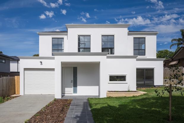 Front exterior view of a contemporary two-storey white rendered residence with a flat roof, strong rectangular geometry, dark-framed windows, an integrated single garage, covered entry porch, paved walkway and front lawn.