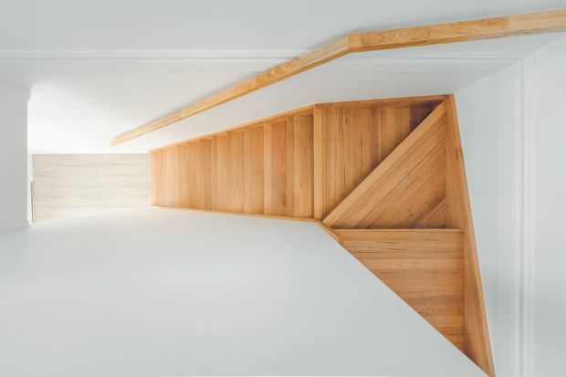 A rotated view of a compact timber staircase with warm natural wood treads, risers, and balustrade detailing, set against white painted walls and ceiling. A simple timber beam runs diagonally overhead, creating a clean, understated stairwell composition.