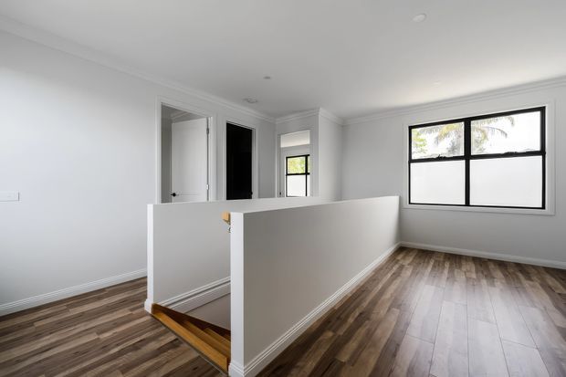 Bright, mostly empty upper-level interior with white walls, decorative cornices, black-framed windows, and warm timber flooring. A half-height partition encloses a stair opening with visible timber steps, while doors and a small window suggest circulation to adjacent rooms.