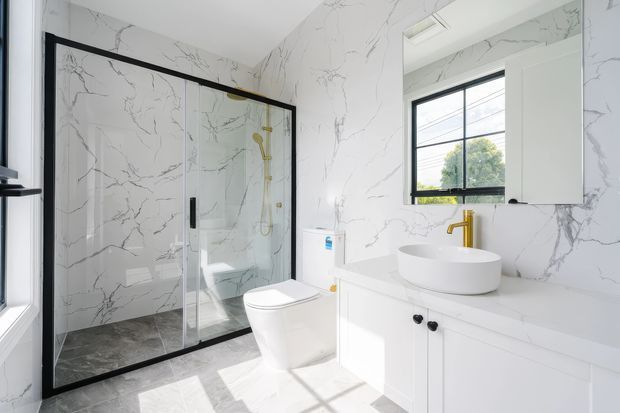 Bright contemporary bathroom with full-height white marble-look wall tiling, a black-framed glass shower enclosure, white toilet, freestanding vessel basin on a white vanity, and a gold-toned tap and shower fittings. A black-framed window brings in natural light, while grey stone-look floor tiles provide contrast.