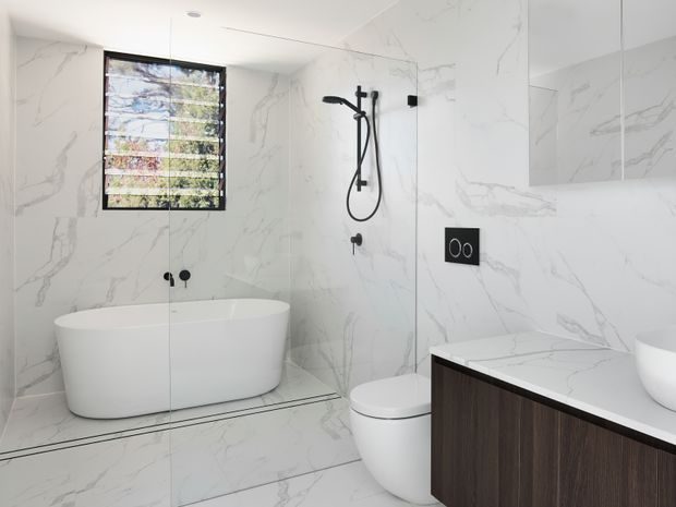 Contemporary bathroom featuring a freestanding white bathtub, walk-in glass shower, wall-hung toilet, and dark timber vanity with a white marble-look surface. Large white marble-look wall and floor tiles with grey veining create a bright, refined finish, complemented by matte black shower and plumbing fixtures. A louvred window provides natural light.