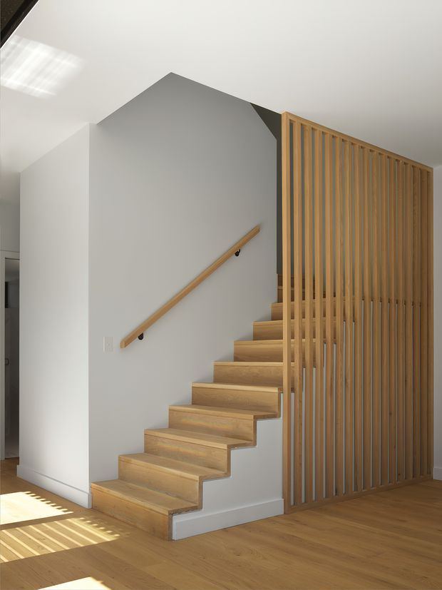 A bright, minimalist interior stairway with open timber treads rising alongside a white wall. A simple timber handrail follows the stair, while a full-height vertical timber slat screen forms the balustrade and partitions the stair from the adjacent space. Warm timber flooring and strong sunlight create a clean, calm atmosphere.