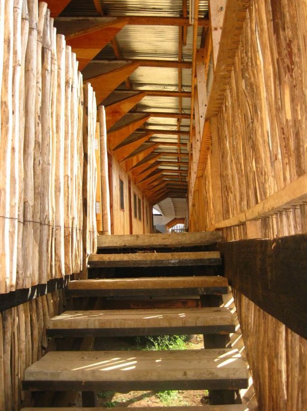 A narrow elevated timber walkway or stair passage constructed from rough natural wood, with open-riser stairs, vertical stick or branch-like wall cladding, exposed timber roof trusses, and corrugated metal roofing. Strong linear perspective leads toward a small opening at the far end, with sunlight filtering through the structure.