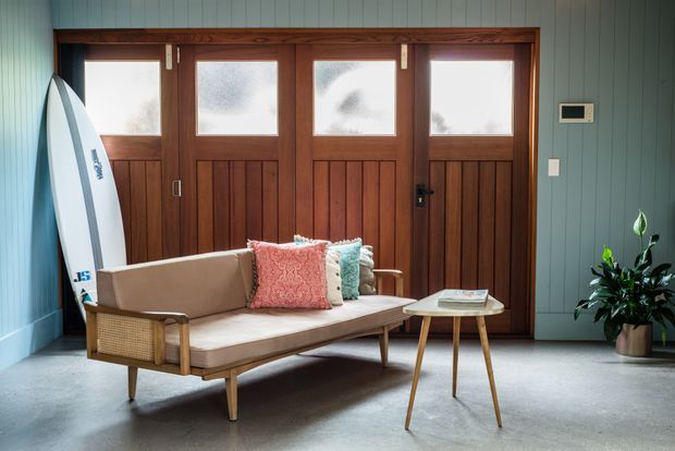 A compact, thoughtfully styled entry or sitting area with a mid-century-inspired timber-framed sofa, woven side panels, patterned and textured cushions, and a small timber side table. The room features blue-green vertical wall panelling, a polished concrete floor, and a row of dark stained timber doors with frosted glass upper panels. A surfboard and potted plant add casual coastal character.